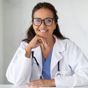Cheerful Mature Doctor Woman Wearing Uniform Smiling To Camera, Sitting At Desk In Clinic Office. Portrait Of Professional Therapist Caucasian Lady Concept. Free Space For Text